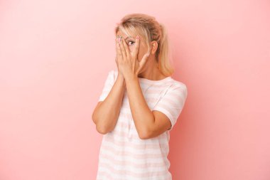 Pembe arka planda izole edilmiş genç Rus kadın parmaklarının arasından korkmuş ve gergin bir şekilde göz kırpıyor..