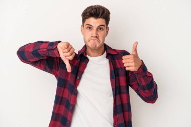 Beyaz arka planda izole edilmiş genç beyaz bir adam baş parmaklarını aşağı ve yukarı kaldırıyor, zor seçim konsepti