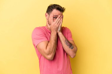 Sarı arka planda izole edilmiş genç beyaz adam parmaklarının arasından korkmuş ve gergin bir şekilde göz kırp..