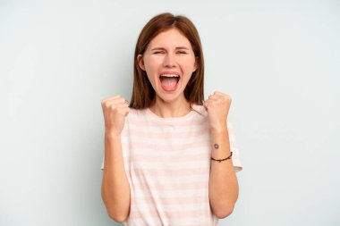 Genç İngiliz kadın mavi arka planda izole edilmiş kaygısız ve heyecanlı tezahüratlar yapıyor. Zafer konsepti.