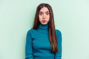 Yeşil arka planda izole edilmiş genç beyaz kadın yanakları patlatıyor, yorgun bir ifadesi var. Yüz ifadesi kavramı.