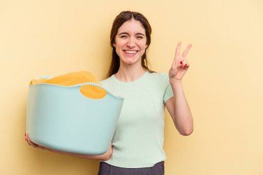 Sarı arka planda izole edilmiş bir sepet tutan beyaz bir kadın neşeli ve tasasız bir şekilde parmaklarıyla barış sembolü gösteriyor..