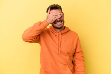 Sarı arka planda izole edilmiş genç beyaz adam elleriyle gözlerini kapatıyor, geniş bir gülümsemeyle bir sürpriz bekliyor..