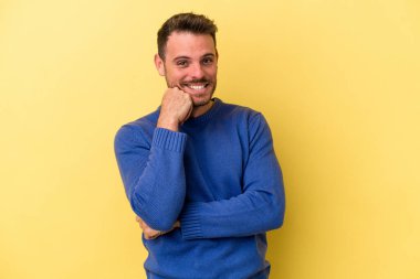 Sarı arka planda izole edilmiş genç, mutlu ve kendinden emin gülümseyen, elleriyle çenesine dokunan beyaz bir adam..