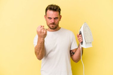 Sarı arka planda demir tutan genç beyaz bir adam kameraya yumruk gösteriyor, agresif yüz ifadesi..