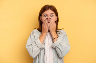 Sarı arka planda izole edilmiş genç bir İngiliz kadın şok olmuş, ağzı ellerle kaplanmış, yeni bir şey keşfetmek için sabırsızlanıyor..
