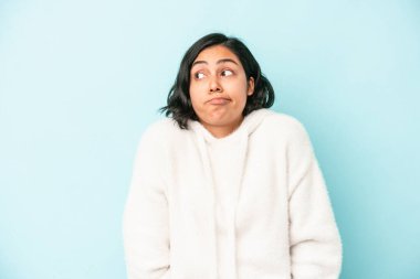 Mavi arka planda izole edilmiş genç Latin kadın omuzlarını silker ve gözleri açık kalır..