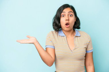 Mavi arka planda izole edilmiş genç Latin kadın, avuç içinde kopya alanı tutmaktan etkilenmiş..