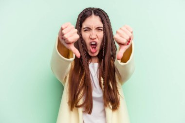 Yeşil arka planda izole edilmiş genç beyaz kadın baş parmağını aşağı gösteriyor ve hoşlanmadığını ifade ediyor..