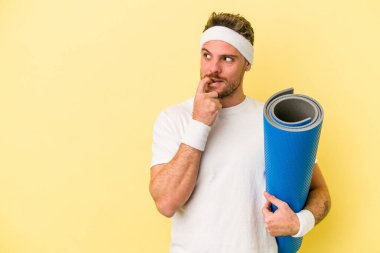 Genç beyaz adam yoga derslerine gidiyor. Elinde sarı arka planda izole edilmiş bir minder var. Bir şeyin fotokopi odasına baktığını düşünüyor..