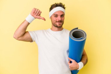 Sarı arka planda izole edilmiş bir minderi tutarken yoga derslerine giden genç beyaz adam gururlu ve kendinden emin hissediyor, örnek olarak..