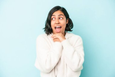 Mavi arka planda izole edilmiş genç Latin bir kadın şans için dua ediyor, şaşırmış ve ağzını açıyor..