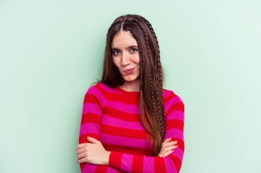 Yeşil arka planda izole edilmiş genç beyaz kadın hoşnutsuzca kaşlarını çatmış, kollarını kavuşturmuş..