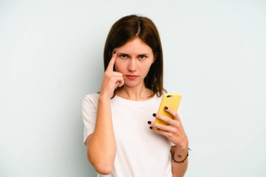 Elinde cep telefonu olan genç bir İngiliz kadın mavi arka planda izole edilmiş. Şakağı işaret ediyor, düşünüyor ve bir göreve odaklanıyor..