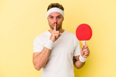 Sarı arka planda pinpon raketi tutan genç beyaz bir adam bir sır saklıyor ya da sessizlik istiyor..