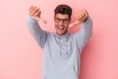 Pembe arka planda izole edilmiş genç beyaz adam iki parmağını da kaldırıyor, gülümsüyor ve kendinden emin..