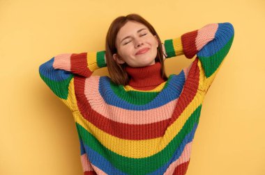 Sarı arka planda izole edilmiş genç bir İngiliz kadın kendini güvende hissediyor, elleri başının arkasında..