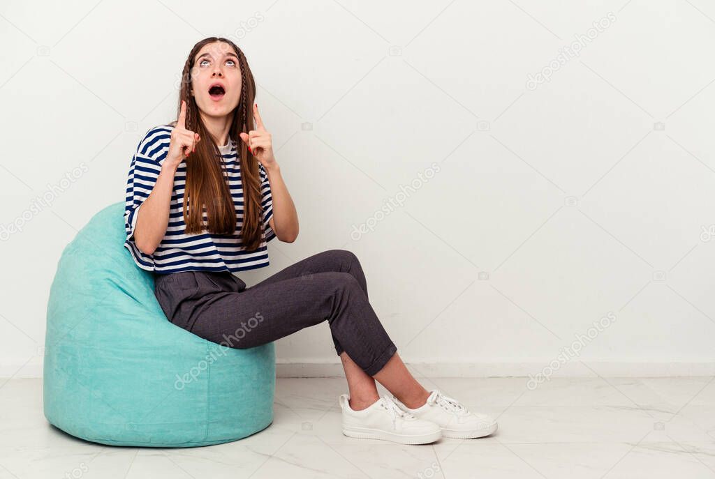 Young caucasian woman sitting on a puff isolated on white background pointing upside with opened mouth.