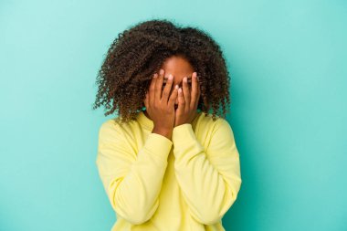 Mavi arka planda izole edilmiş kıvırcık saçlı, genç Afro-Amerikalı kadın kameraya parmaklarıyla göz kırpıyor, yüzünü kapatmaktan utanıyor..