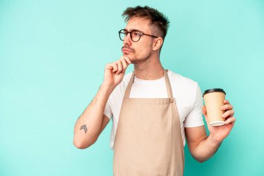 Genç tezgahtar elinde mavi arka planda izole edilmiş bir kahve tutarken şüpheli ve şüpheci bir ifadeyle yana bakıyor..