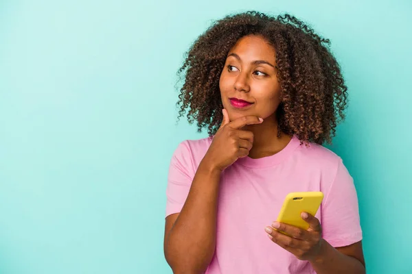 Młoda Afroamerykanka Trzymająca Telefon Komórkowy Niebieskim Tle Patrząca Bokiem Wątpliwym — Zdjęcie stockowe
