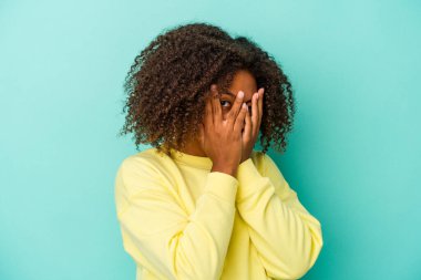Mavi arka planda izole edilmiş kıvırcık saçlı, genç Afro-Amerikalı kadın parmaklarının arasından korkmuş ve gergin bir şekilde göz kırpıyor..