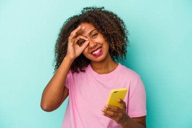 Genç Afro-Amerikalı bir kadın elinde cep telefonu ile mavi arka planda izole edilmiş bir şekilde heyecanlı..