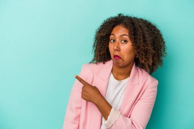 Mavi arka planda izole edilmiş kıvırcık saçlı, genç Afro-Amerikalı kadın gülümseyip işaret ediyor, boş alanda bir şey gösteriyor..