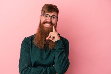Uzun sakallı beyaz bir adam pembe arka planda izole edilmiş mutlu ve kendinden emin gülümsüyor, çenesine elleriyle dokunuyor..