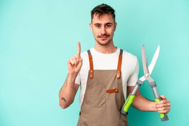 Genç bahçıvan, elinde makas tutan beyaz adam mavi arka planda bir numarayı parmağıyla gösteriyor..