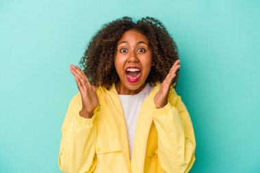 Mavi arka planda izole edilmiş kıvırcık saçlı genç Afro-Amerikalı kadın zaferi veya başarıyı kutluyor, şaşırıyor ve şok oluyor..