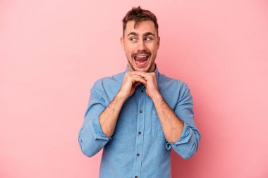 Pembe arka planda izole edilmiş genç beyaz adam ellerini çenesinin altında tutuyor, mutlu bir şekilde yana bakıyor..