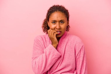 Pembe arka planda izole edilmiş, tırnak yiyen, gergin ve endişeli bornoz giyen genç Afrikalı Amerikalı kadın..