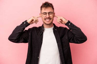 Pembe arka planda izole edilmiş genç beyaz bir adam parmaklarıyla kulaklarını kapatıyor, gürültülü bir ortam yüzünden stresli ve çaresiz..