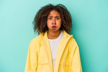 Mavi arka planda izole edilmiş kıvırcık saçlı, genç Afro-Amerikalı kadın yanakları siliyor, yorgun bir ifadeye sahip. Yüz ifadesi kavramı.
