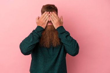 Uzun sakallı, beyaz, genç bir adam pembe arka planda izole edilmiş. Gözleri elleriyle kapamaktan korkuyor..