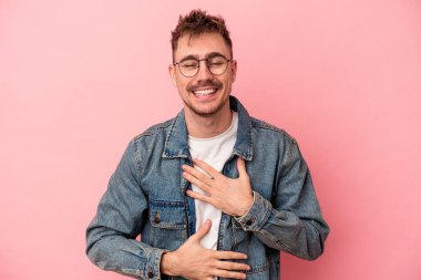 Pembe arka planda izole edilmiş genç beyaz adam mutlu bir şekilde gülüyor ve ellerini karnının üstünde tutarak eğleniyor..