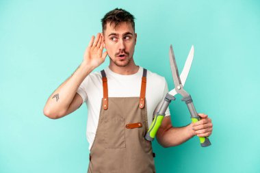 Genç bahçıvan Kafkasyalı bir adam mavi arka planda izole edilmiş bir makas tutuyor ve bir dedikoduyu dinlemeye çalışıyor..