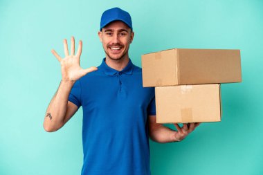 Genç teslimatçı beyaz adam mavi arka planda izole edilmiş. Gülümsüyor beş numarayı parmaklarıyla gösteriyor..