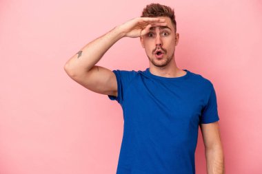 Pembe arka planda izole edilmiş genç beyaz adam uzaklara bakıyor ve elini alnına koyuyor..