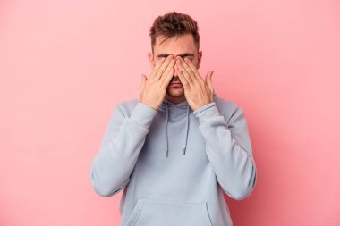 Pembe arka planda izole edilmiş genç beyaz adam elleriyle gözlerini kapatmaktan korkuyor..