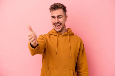 Pembe arka planda izole edilmiş genç beyaz adam selamlama jesti olarak kameraya elini uzatıyor..