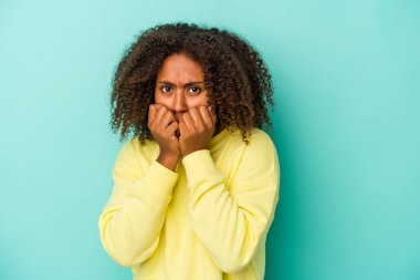 Kıvırcık saçlı, mavi arka planda, tırnak yiyen, gergin ve endişeli genç Afro-Amerikan kadın..
