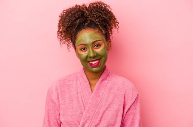 Bornoz ve yüz maskesi takan genç Afrikalı Amerikalı kadın pembe arka planda izole edilmiş mutlu, gülümseyen ve neşeli..
