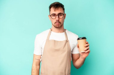 Genç tezgahtar elinde mavi arka planda izole edilmiş bir kahve tutarken omuzlarını silker ve gözleri açık kalır..
