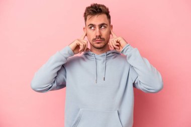 Pembe arka planda izole edilmiş genç beyaz bir adam parmaklarıyla kulaklarını kapatıyor, gürültülü bir ortam yüzünden stresli ve çaresiz..