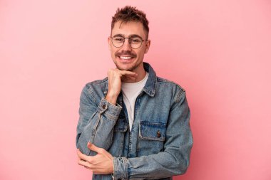 Pembe arka planda izole edilmiş genç beyaz adam gülümsüyor ve kendinden emin, elleriyle çenesine dokunuyor..