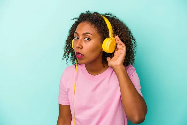 Young african american woman listening to music isolated on blue background trying to listening a gossip.