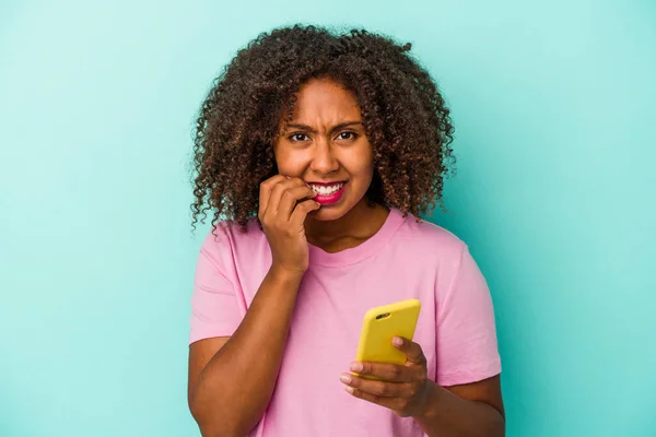 Młoda Afroamerykanka Trzymająca Telefon Komórkowy Niebieskim Tle Obgryzająca Paznokcie Nerwowa — Zdjęcie stockowe