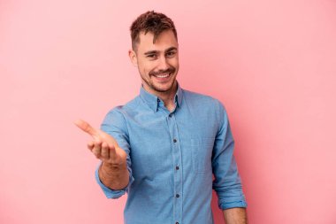Pembe arka planda izole edilmiş genç beyaz adam selamlama jesti olarak kameraya elini uzatıyor..